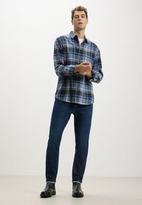 Camicia di flanella a quadri navy, grigia e blu chiaro; maniche lunghe; davanti con bottoni. Abbinata a jeans in denim blu scuro e stivali in pelle nera.