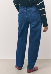 Zilas džinsa bikses ar brīvu piegriezumu, ar divām aizmugurējām kabatām un taisnu kāju dizainu. Valkātas ar sarkaniem apaviem un melnu svītrainu džemperi.