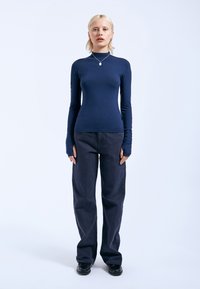 Dunkelblaues Langarmshirt mit hohem Kragen, getragen mit locker sitzenden dunklen Hosen und schwarzen Schuhen. Der Stoff wirkt glatt; die Ärmel haben Daumenlöcher.