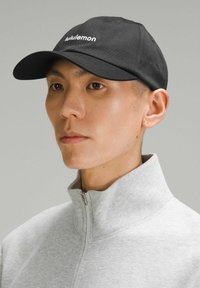 Crna kapa s zakrivljenim vizirjem, s bijelim natpisom "lululemon". Nosena s svijetlosivim zip-up kragnom. Glatka tkanina.