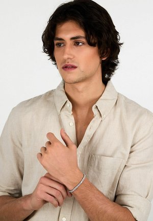 Bracciale d'argento con accenti rettangolari su una camicia di lino beige a vestibilità comoda, indossato su un polso maschile. La pelle è leggermente abbronzata e i capelli sono di lunghezza media.