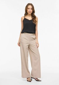 Camisole en satin noir avec bordure en dentelle, associée à un pantalon beige à jambes larges et taille haute. Le mannequin pose devant un fond blanc uni.