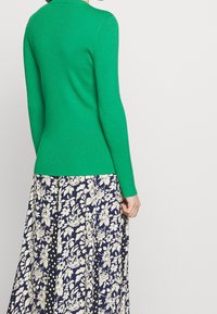 Vihreä ribbineulottu pitkähihaisen neulepaidat, yhdistetty kukikkaaseen, tummansiniseen hameeseen, jossa on kermaisia kuvioita ja mustia yksityiskohtia.