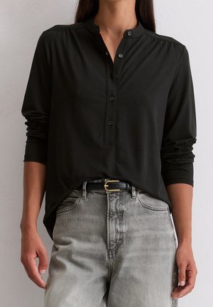 Femme portant une chemise noire à manches longues boutonnée rentrée dans un jean gris clair délavé avec une ceinture noire à boucle dorée.