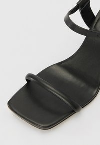 Musta sandaali, jossa on neliömäinen varvasosa ja ohuet nauhat. Silkkinen nahkamateriaali mattapinnalla. Minimalistinen muotoilu ilman näkyviä metalliosia.