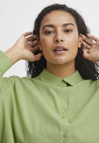 Camicia verde chiaro con bottone e colletto, realizzata in tessuto morbido e dal taglio morbido. Il modello ha i capelli ricci e le mani vicino al viso.