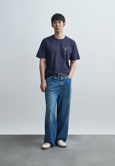 Μπλε βαμβακερό μπλουζάκι με ένα ανοιχτό σχέδιο στο αριστερό στήθος, σε συνδυασμό με φαρδιά μπλε τζιν παντελόνια και λευκά αθλητικά παπούτσια πάνω σε μια υφή επιφάνεια.