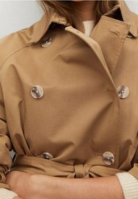 Trench-coat beige croisé avec de gros boutons gris clair. Il présente une taille ceinturée et une texture de tissu lisse.