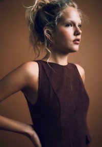 Top marrone a costine senza maniche con un collo alto e cuciture sottili, abbinato a orecchini pendenti in gemma rossa su uno sfondo caldo.