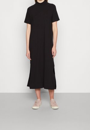 Persona que lleva un vestido midi negro sencillo de manga corta con aberturas laterales y zapatillas casual color rosa claro, de pie contra una pared blanca.
