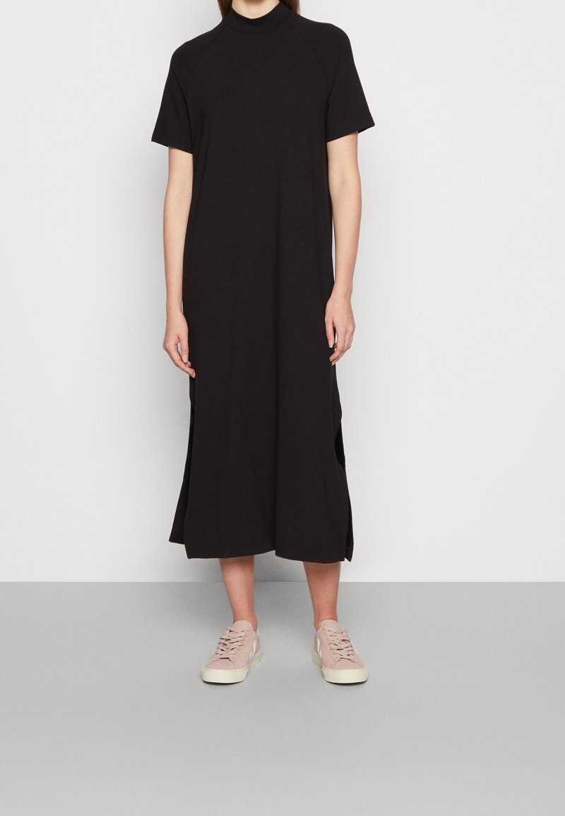 Personne portant une robe midi noire unie à manches courtes avec des fentes latérales et des baskets décontractées rose clair, debout contre un mur blanc.
