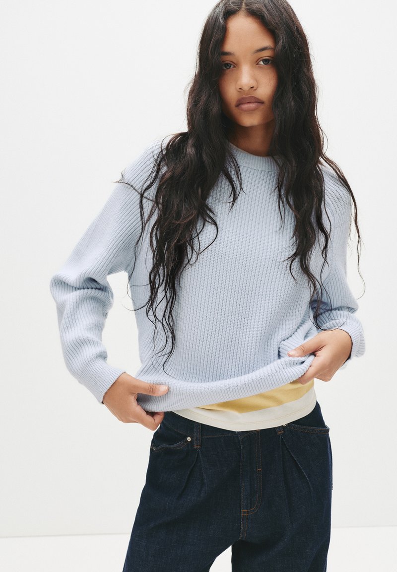 Maglione azzurro chiaro a coste con colletto a girocollo, abbinato a pantaloni in denim scuro. Il maglione presenta maniche lunghe e una vestibilità comoda.