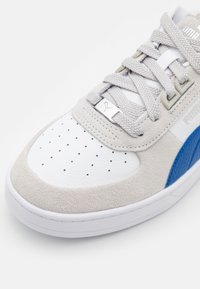 Ljusgrå mocka- och vitlädersneaker med blå detaljer, perforerad tå och grå skosnören. Har märkta silverdetaljer och texturerade inslag.