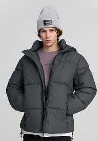 Ciemnoszara kurtka puchowa z wysokim kołnierzem, pikowaną fakturą i zamkiem z przodu. Noszona na jasnofioletowej koszuli, z jasnoszarym beanie.