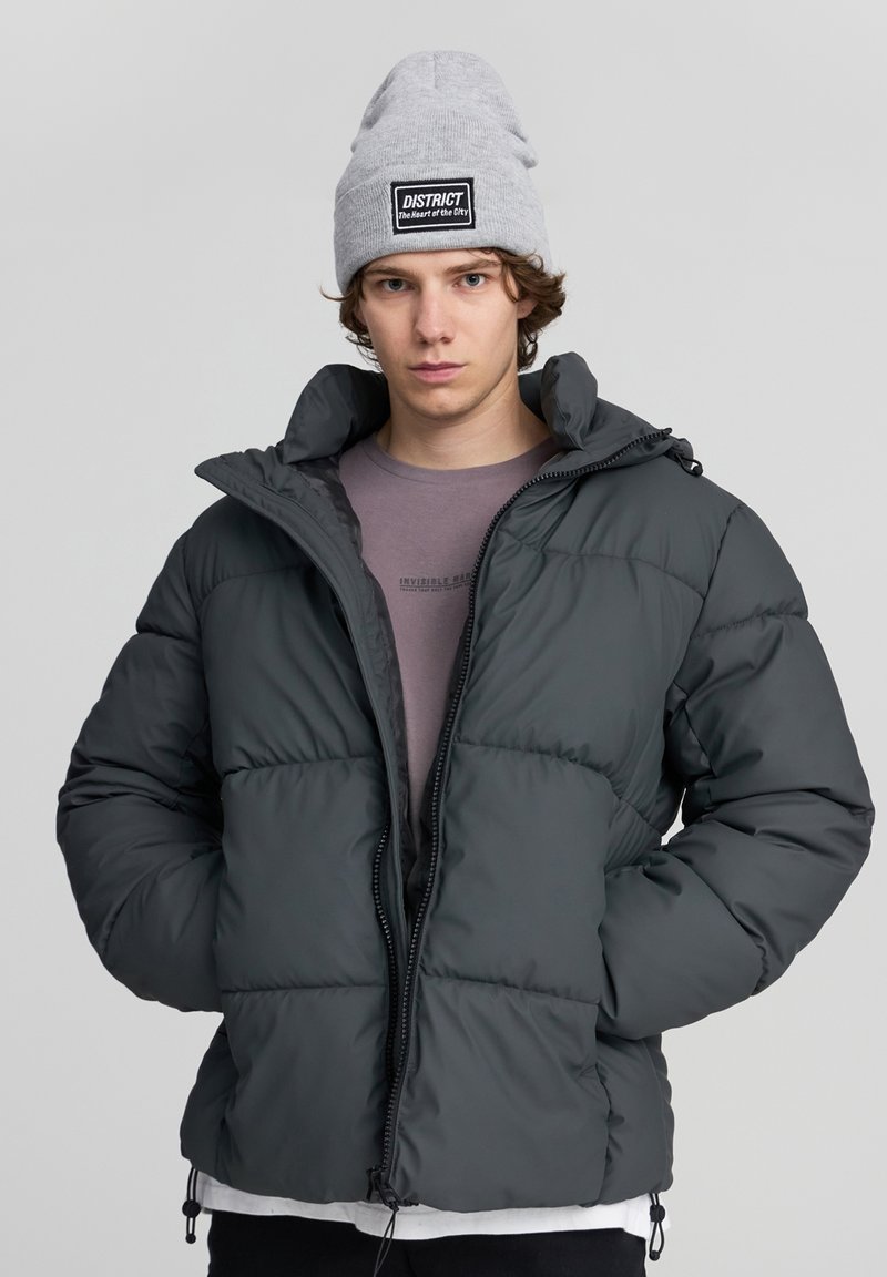 Ciemnoszara kurtka puchowa z wysokim kołnierzem, pikowaną fakturą i zamkiem z przodu. Noszona na jasnofioletowej koszuli, z jasnoszarym beanie.