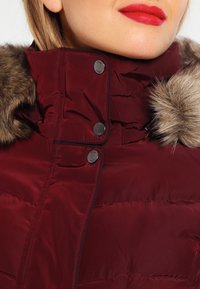 Vinröd vadderad jacka med hög krage, prydd med konstpäls och metalliska tryckknappar. Slät textur och quiltad design.