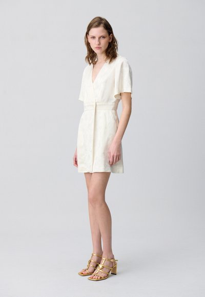 Robe blanche courte avec un col en V, des manches courtes et une taille ajustée. Tissu texturé avec un motif subtil. Associée à des sandales à talons dorées et à lanières.