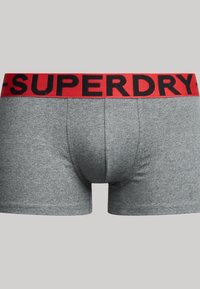 Boxer shorts en coton gris avec une large ceinture élastique rouge arborant le lettrage noir "SUPERDRY." Design à jambes courtes, texture lisse.
