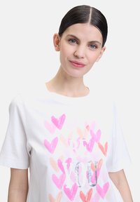 T-shirt en coton blanc avec des motifs de cœurs roses et pêches et le texte "BE YOU" en lettres argentées. Manches courtes et coupe décontractée.