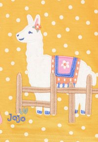 Sárga szövet fehér pöttyökkel, rajta egy hímzett függőleges fehér lámával, mintás nyereggel, virágos díszítéssel és egy barna kerítéssel.