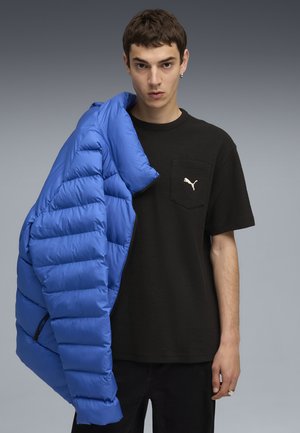 Padded blue jacket with quilted texture held over a black short-sleeve shirt with a chest pocket and a small Puma logo. Black pants.