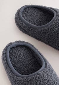 Graue Plüsch-Hausschuhe mit abgerundeten Öffnungen, einer strukturierten Außenseite und einem glatten Stoffbesatz um die Öffnung.