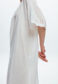 Robe blanche en tissu léger, avec des manches courtes ornées de broderies à œillets et d'un bord festonné, d'une coupe décontractée et de fentes sur les côtés.