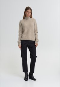 Bžūp megztinis šviesios spalvos su aukštu kaklu, derinamas su juodomis tiesioms kojoms kelnėmis ir juodais batais iki kulkšnių, fonas neutralaus atspalvio.