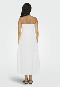 Vestido maxi blanco sin tirantes con un corpiño fruncido y una falda fluida. La parte posterior presenta un detalle elástico, combinado con sandalias negras.