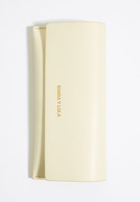Cartera de imitación cuero color crema con una textura suave, que presenta un logo estampado en dorado y un diseño minimalista de cierre tipo folio.
