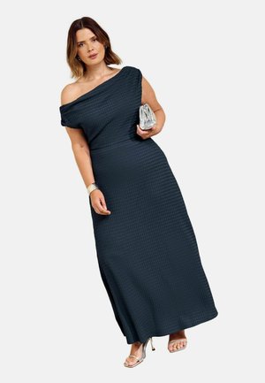 Kvinde iført en tekstureret mørkeblå en-øjet lång kjole, der holder en sølv clutch og bærer sølv åbne hæle og et armbånd.