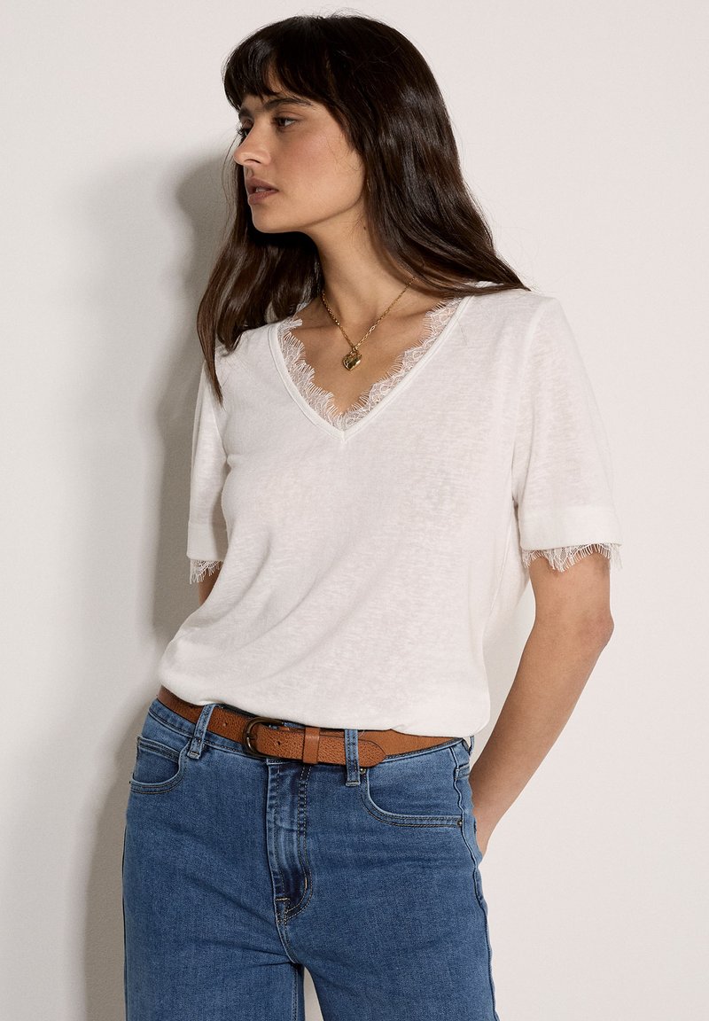 T-shirt blanc à manches courtes avec un col en V, orné de dentelle le long du col et des manches. Associé à un jean bleu et une ceinture marron.