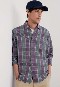 Camisa de cuadros de manga larga en un patr�ón de cuadros en verde, púrpura y blanco. Cuello tipo button-down y bolsillo frontal, combinada con una gorra de béisbol azul marino.