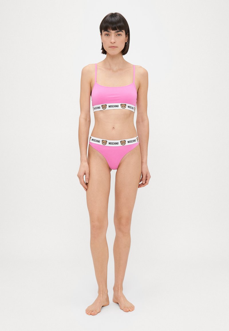 Roza dvodelna komplet, ki vključuje braletko s tankimi naramnicami in bikini hlačke, oba z kontrastnim belim pasom z oznako "Moschino."