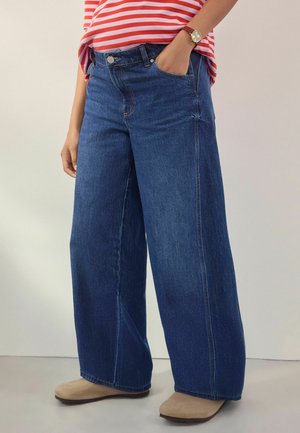 Hellebeine Jeans aus dunklem Denim mit hohem Bund, fünf Taschen und subtiler Textur. Kombiniert mit beigen Schuhen.