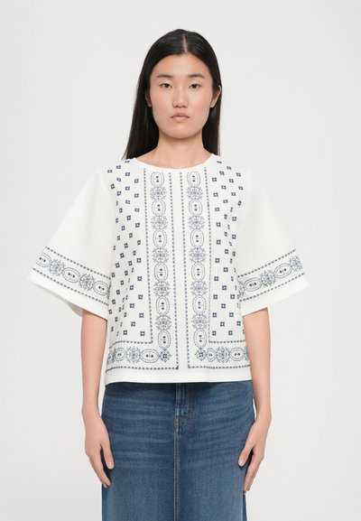 Haut blanc à manches courtes avec de délicates broderies bleu marine, présentant des motifs géométriques et floraux le long de l'encolure et de l'ourlet.