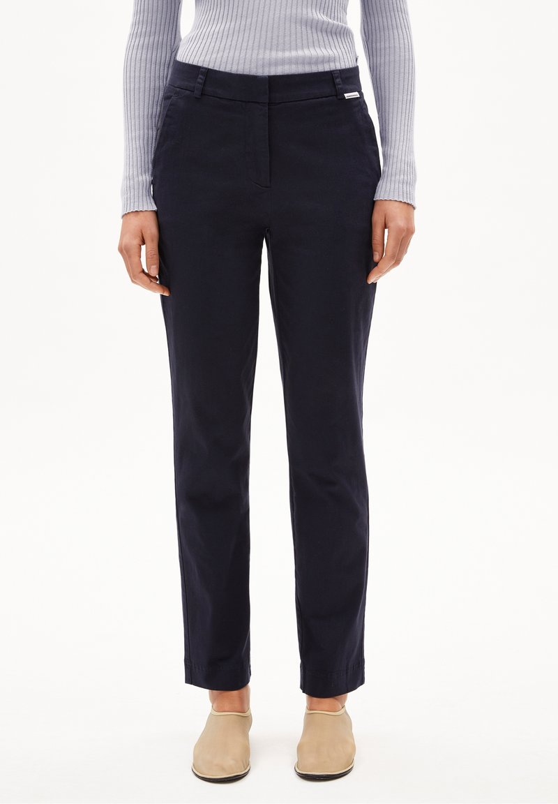 Femme portant un pantalon noir à jambes droites, un haut gris clair côtelé à manches longues et des chaussures beige à enfiler sur fond blanc.