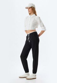 Witte cropped shirt met een kraag, zwarte joggers met een trekkoord in de taille en witte sneakers met lichtbruine zolen. Lichtgrijze pet op het hoofd.