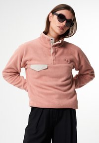 Hellrosa Fleece-Pullover mit hohem Kragen, knöpfbarer Leiste und kontrastierendem beigen Brusttasche. Weicher Stoff, lässige Passform.