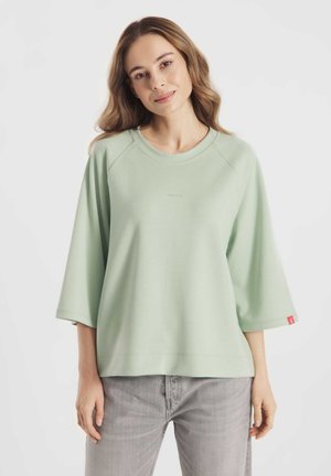 Vrouw met lang golvend haar, gekleed in een lichtgroene oversized trui en grijze spijkerbroek, staand tegen een eenvoudige witte achtergrond.