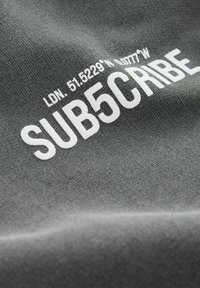 Tissu gris avec du texte imprimé blanc incluant des coordonnées géographiques et le mot "SUB5CRIBE", mettant en avant une texture lisse.