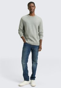 Grijze gebreide trui, blauwe spijkerbroek en witte sneakers. De trui heeft een ronde hals en geribbelde boorden. De spijkerbroek heeft een klassieke pasvorm.