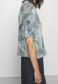Blusa a maniche corte in un motivo turchese, blu navy e bianco. Presenta una vestibilità morbida e dettagli arricciati sulle maniche. Tessuto leggero.