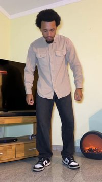 Camicia button-up marrone con due tasche sul petto, jeans neri e sneakers nere con accenti bianchi. In piedi in una stanza con una TV e un riscaldatore decorativo.