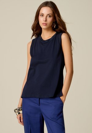 Giovane donna con lunghi capelli castani, indossa una canotta blu navy, pantaloni blu, orecchini geometrici e tiene dei braccialetti con una mano in tasca.
