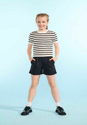 Smiling girl stands with hands in pockets wearing black shorts, black-and-white striped short sleeve top, white socks, and black shoes on blue background.