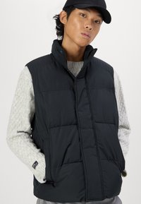 Vesta puffer neagră cu guler înalt, închidere cu fermoar și buzunare laterale, purtată peste un pulover texturat gri deschis. Design minimalist.