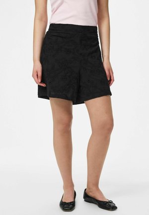 Femme portant un short noir à texture florale, un haut rose sans manches et des ballerines noires, debout sur un fond blanc.