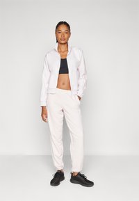 Lichtroze zip-up jacket met ribbelcuff, gecombineerd met bijpassende joggers; een zwarte sport-bh en zwarte sneakers maken de outfit compleet.