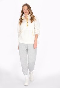Crème fleece pullover met gele accenten, halve knoopsluiting en een voorkant zak; gecombineerd met grijze joggingsbroek en witte sneakers.